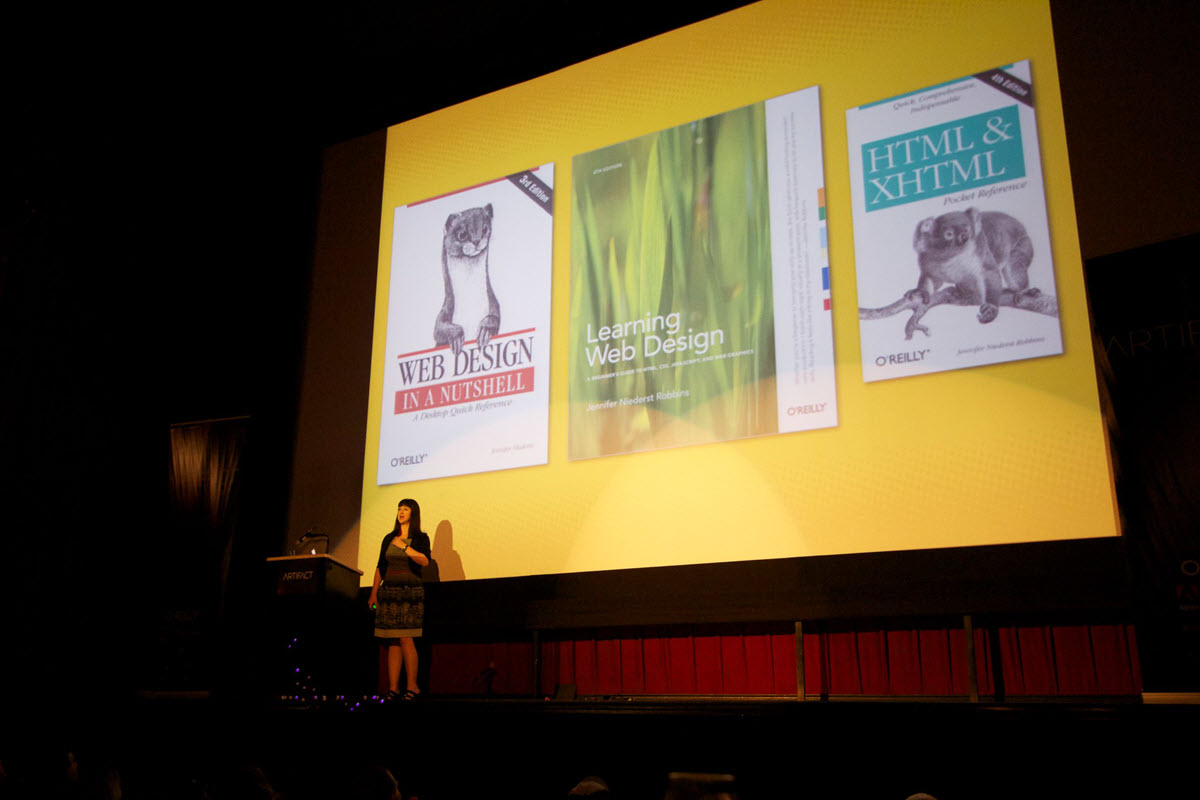 A speaker on stage with photos of books behind her